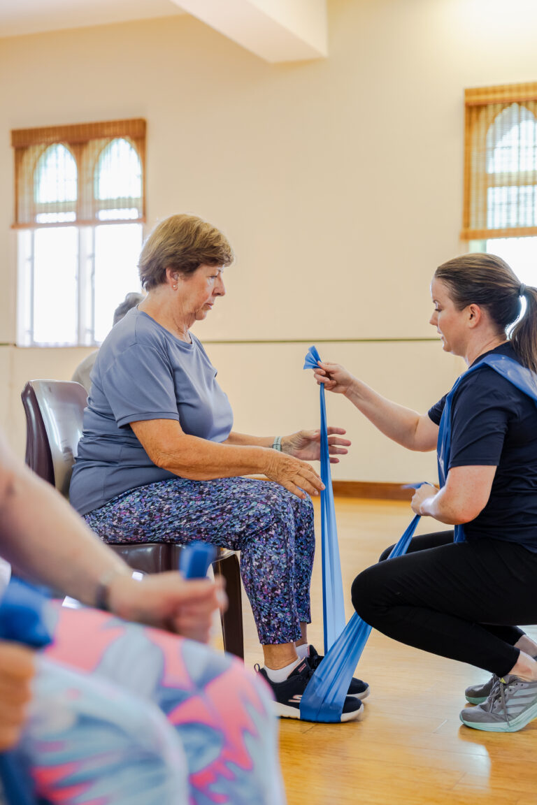 An instructor helps an older woman use a resistance band during a seated exercise.