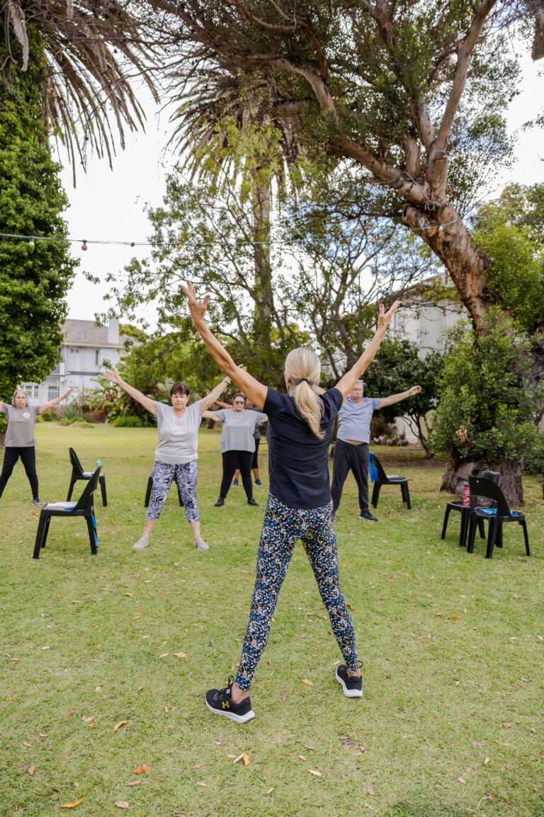 Instructor leading people over 60 through standing arm raises outdoors in a group.