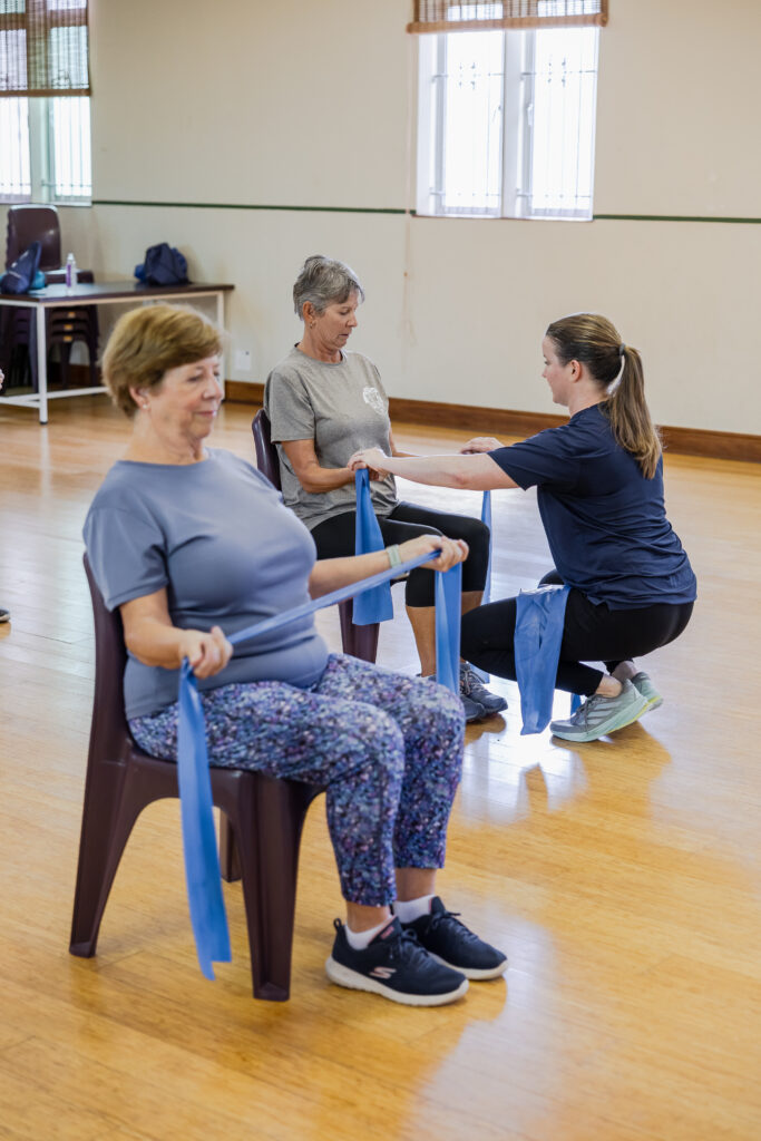 People over 60 practising seated resistance-band exercises while an instructor provides hands-on guidance.