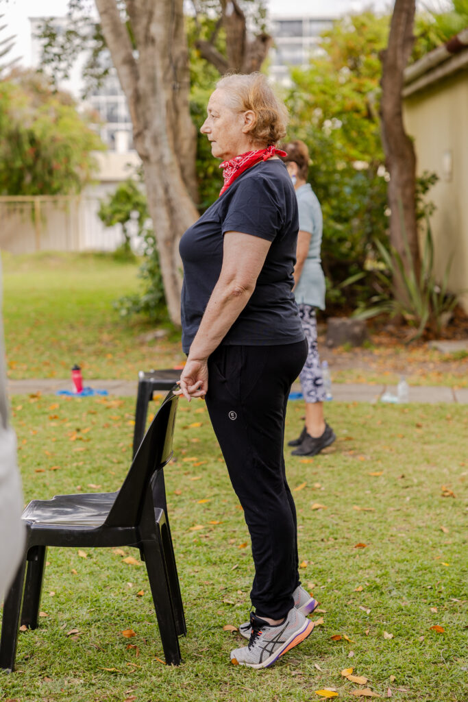 Person over 60 practising a supported standing balance exercise using a chair outdoors.